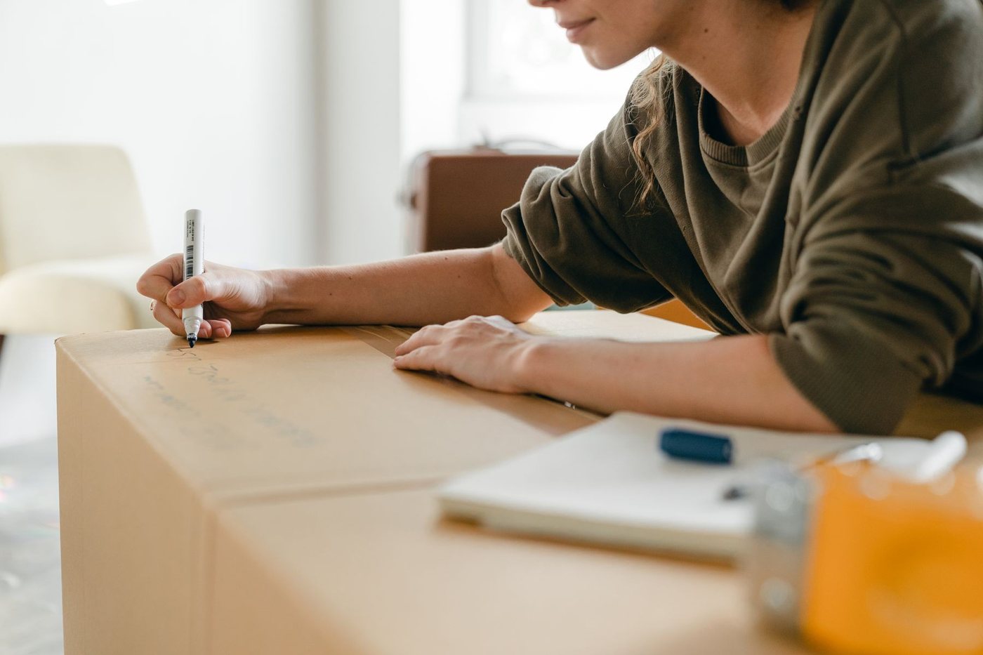 A hand writing a room label on a packed carton, notebook and marker resting on the lid.